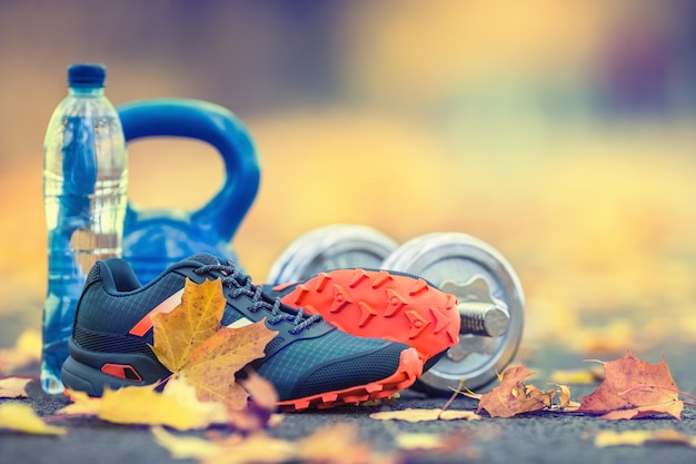 Paire de chaussures de sport bleu eau et haltères posées sur un chemin dans une allée d'automne avec des feuilles d'érable