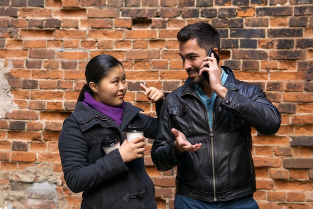 Paire de jeunes sur fond de mur, le gars parle simultanément avec la fille et au téléphone