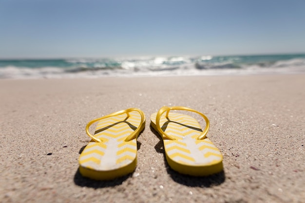 Paire de tongs à motifs en zigzag jaune et blanc sur une plage de sable avec une mer calme et un ciel bleu clair en été