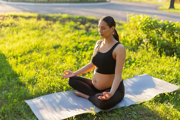 Paisible jeune femme enceinte positive en costume de gymnastique fait du yoga et médite assis sur un tapis sur