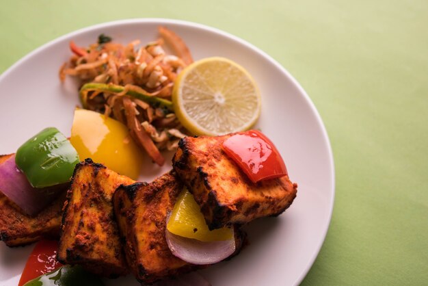 Paneer Tikka ou chili paneer Kabab - Brochettes de fromage indien Tandoori, servies dans une assiette blanche avec poivrons et oignons colorés, avec sauce verte