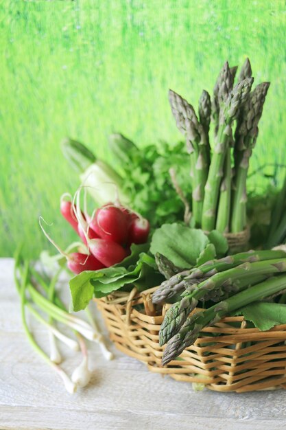 Un panier avec des asperges vertes fraîches, des radis et du persil sur une table en bois.