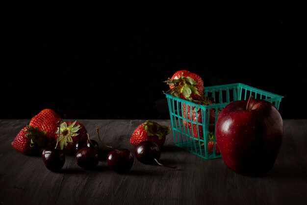 Panier avec des fraises et une pomme avec des cerises sur le côté sur une table en bois vintage noir