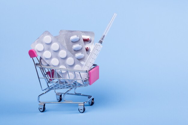 Panier et médicaments. Un chariot avec des blisters de pilules et une seringue sur fond bleu.