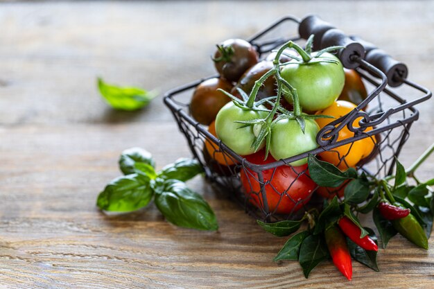 Panier en métal avec différentes tomates rouges, jaunes et vertes mûres sur une table en bois et des feuilles de basilic