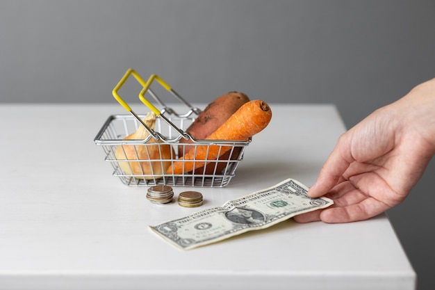 Un panier de nourriture de supermarché en métal avec des légumes et du papier-monnaie et des pièces sur une table blanche