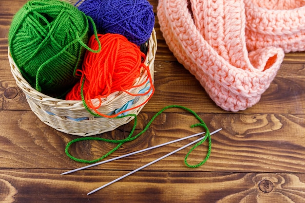 Panier en osier avec fils à tricoter colorés, aiguilles à tricoter et écharpe tricotée sur table en bois