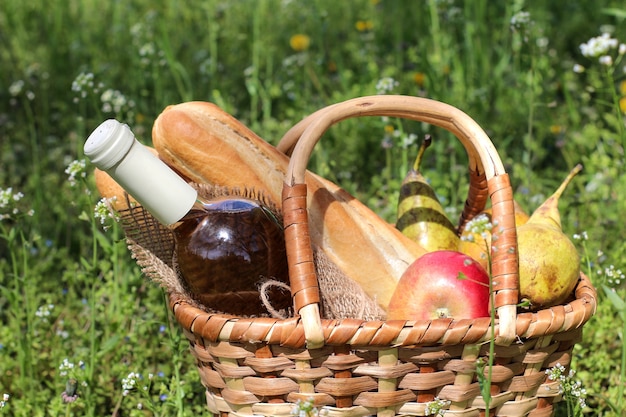 Panier en osier de pique-nique avec du vin, des fruits et d'autres produits dans l'herbe verte.