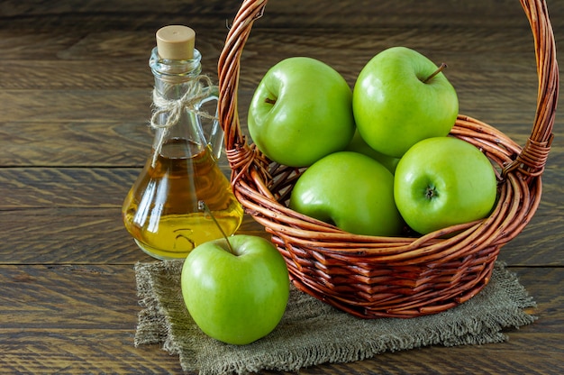 Panier en osier avec pommes vertes mûres et bouteille de vinaigre d'aplle sur fond de bois.