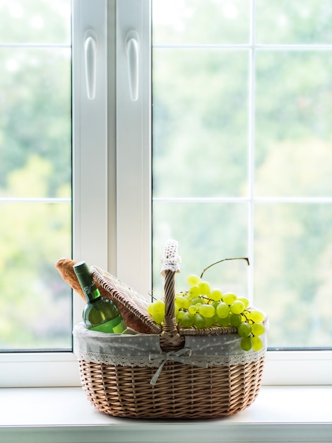 Panier en osier pour pique-nique avec vin, baguette fraîche et raisins debout sur le rebord de la fenêtre