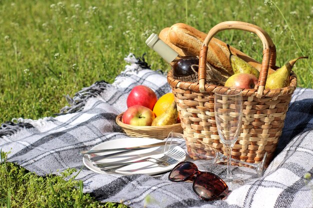 Panier pique-nique avec vin, fruits et autres produits sur une couverture à motifs dans l'herbe.