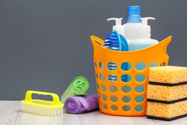 Photo panier en plastique avec bouteilles de liquide vaisselle, nettoyant pour carrelage, détergent pour fours à micro-ondes et cuisinières. brosse, sacs poubelles et éponges sur fond bois et gris. concept de lavage et de nettoyage.