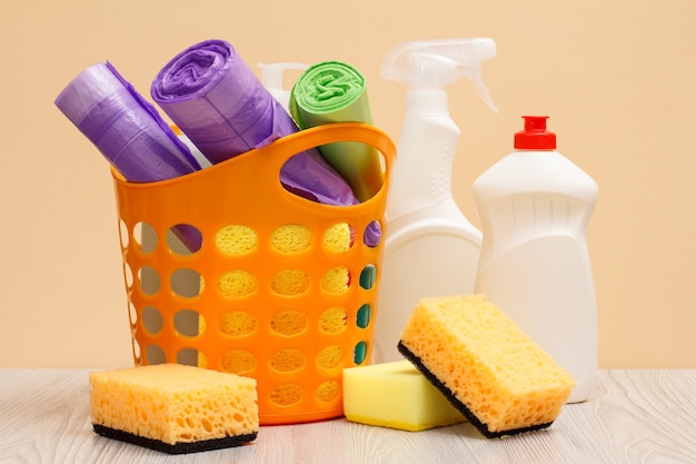 Panier en plastique avec sacs poubelles, bouteilles blanches de nettoyant pour vitres et carreaux, éponges sur fond beige. Concept de lavage et de nettoyage.