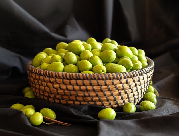 Panier rond en osier rempli d'olives vertes Autour du panier il y a aussi des olives Le fond est en tissu noir