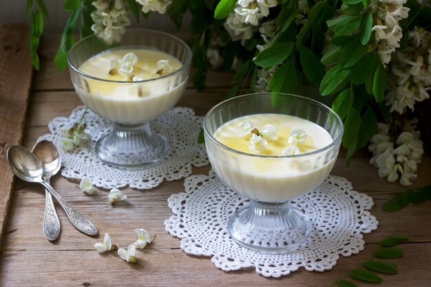 Panna cotta au goût de robinier, servie avec du miel et des fleurs de Robinia blanches. Style rustique.