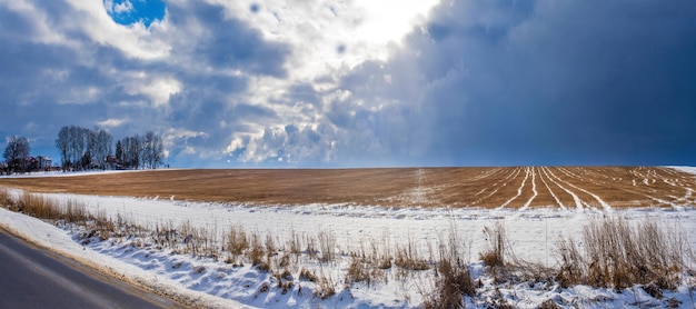 Panorama d'un champ enneigé avec des motifs de chaume après l'avoir récolté près de la route un village et des arbres sous un beau ciel sombre à l'horizon