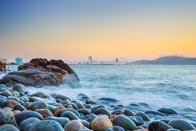 Panorama de la ville de Quy Nhon depuis la plage de galets et les rochers au premier plan, célèbre destination touristique au Vietnam. Quy Nhon bay mer douce vagues longue exposition. gratte-ciel et toits de la ville au coucher du soleil