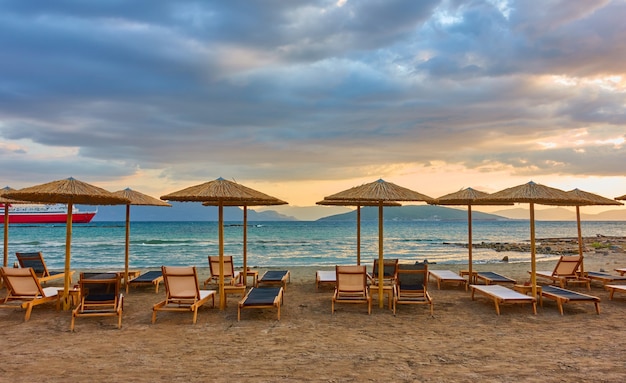 Panorana de plage vide avec chaises longues et parasols en paille au bord de la mer et nuages colorés au coucher du soleil, île d'Egine, Grèce - paysage grec - paysage marin