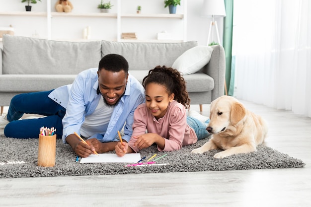 Papa afro-américain aimant dessinant une photo avec sa fille souriante dans le salon, famille heureuse allongée sur un tapis de sol avec Labrador et utilisant des crayons colorés, s'amusant ensemble à la maison