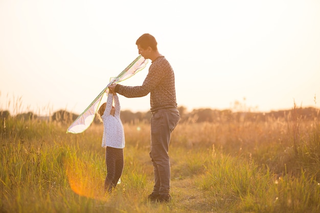 Papa aide sa fille à faire voler un cerf-volant dans un champ en été au coucher du soleil. Animations familiales en plein air, fête des pères, fête des enfants. Milieu rural, accompagnement, entraide. Lumière orange du soleil