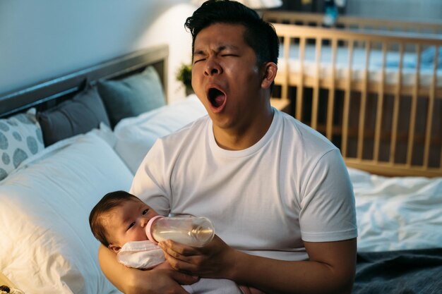 un papa asiatique fatigué pour la première fois est en train de bâiller tout en étant assis à son chevet en train de nourrir sa fille nouveau-née en bouteille de lait tard dans la nuit dans la chambre à la maison.