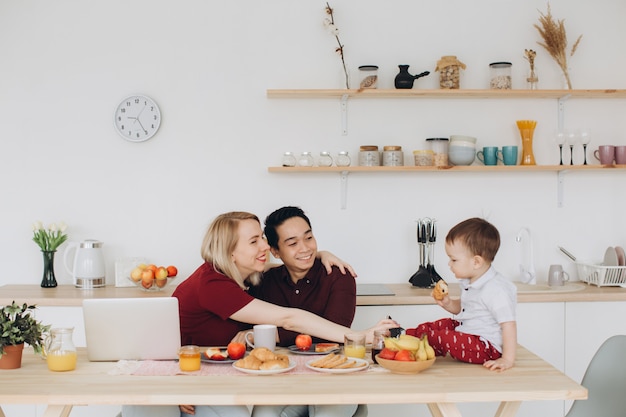 Papa asiatique et maman européenne travaillent avec leurs gadgets, et le fils prend le petit déjeuner seul. Problèmes technologiques modernes.