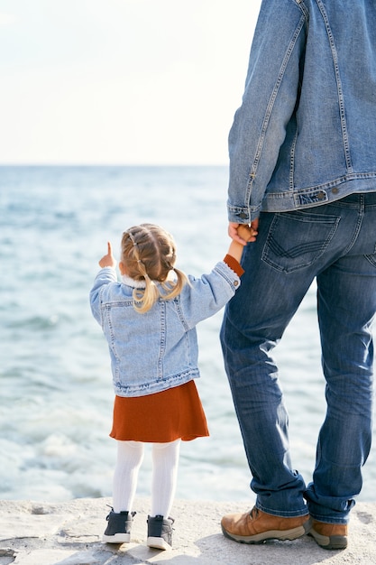 Papa dans une veste en jean et un jean tient la main d'une petite fille qui pointe vers la mer debout