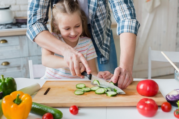 Papa enseigne à sa petite fille à couper le concombre avec un couteau sur une planche à découper, copiez l'espace