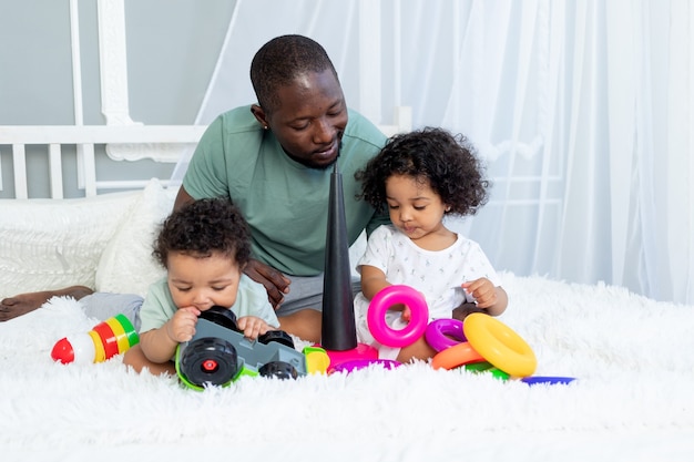 Papa de famille afro-américain avec des bébés enfants joue et collectionne une pyramide colorée à la maison sur le lit, famille heureuse