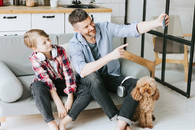 papa de famille jeune homme et fils adolescent assis sur un canapé dans un appartement confortable avec un chien caniche et faisant du selfie au téléphone du temps de qualité avec votre famille