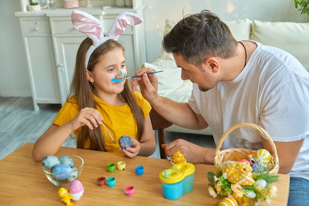 Papa et fille se tachent le visage avec de la peinture bleue pour peindre des œufs. sur la table se trouve un panier avec des œufs de Pâques et des peintures.