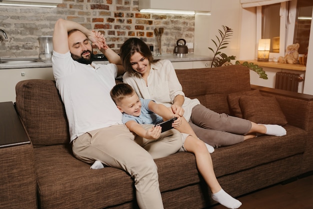 Un papa, un fils et une maman sur le canapé. Un enfant heureux profite du jeu sur le smartphone près de la mère et du père le soir dans une maison confortable.