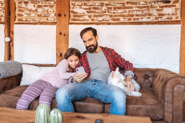 Papa heureux avec sa petite fille regardant le téléphone à la maison assis dans le canapé avec le chien