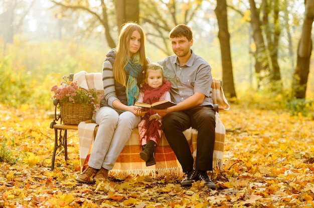 Papa, maman et leur fille sont assis sur un banc dans le parc en automne. Les parents avec une petite fille se sont réfugiés dans une couverture pour se réchauffer.
