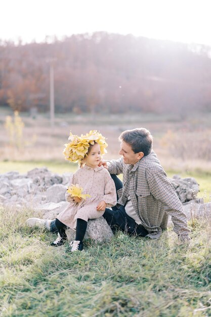 Papa a posé sa main sur l'épaule d'une petite fille assise sur une pierre sur la pelouse et portant une couronne de