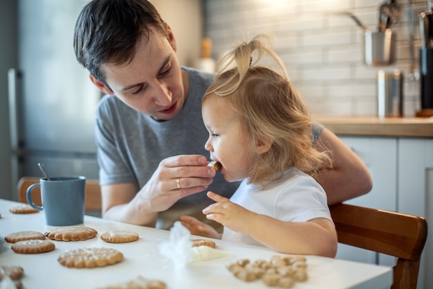 Papa et sa fille décorent le pain d'épice de Noël à la maison un garçon et une fille peignent avec des cornets avec sug...