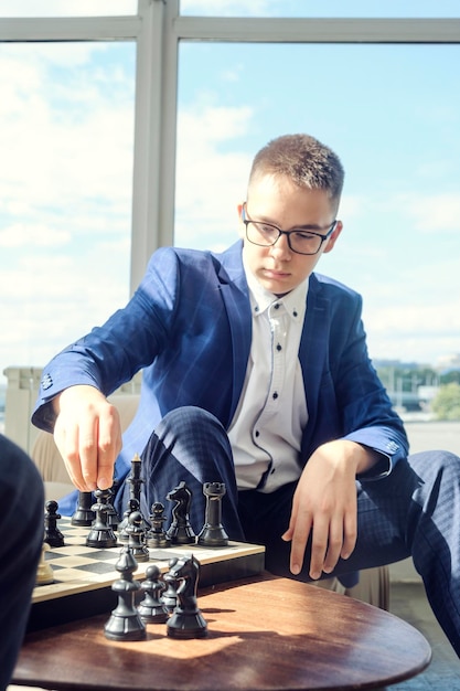 Papa et son fils sont assis à une table près d'une grande fenêtre et jouent aux échecs en faisant des mouvements avec des pièces