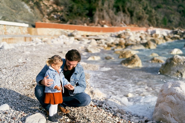 Papa souriant s'est accroupi à côté de la petite fille qu'elle lui donne des cailloux d'une plage rocheuse par le