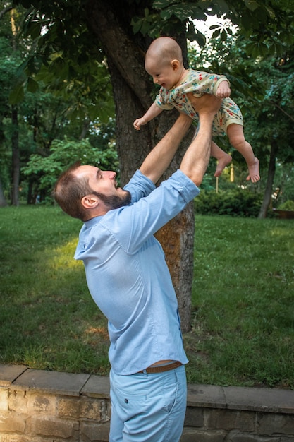 Papa tient son bébé et joue avec lui dans le contexte d'un paysage estival. Bébé sourit et regarde papa