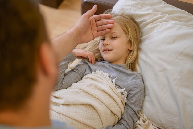 Papa touche le front de son enfant malade allongé sur le canapé recouvert d'une couverture blanche