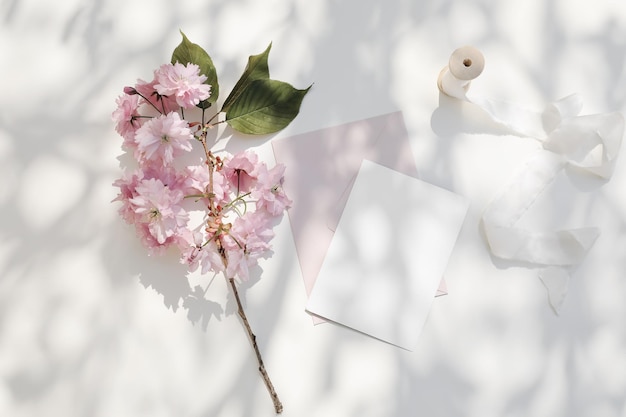 Papeterie de mariage de printemps Branche de sakura de cerisier japonais rose en fleurs au soleil Maquette d'invitation de carte de voeux vierge Enveloppe rose fard à joues Superposition d'ombres de table blanches Plateau plat