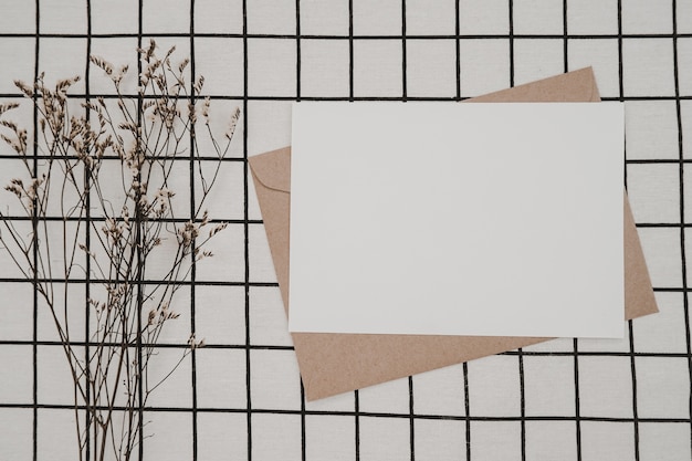 Papier blanc vierge sur enveloppe en papier brun avec fleur sèche Limonium et boîte en carton sur tissu blanc avec motif quadrillé noir. Maquette de carte de voeux vierge horizontale.