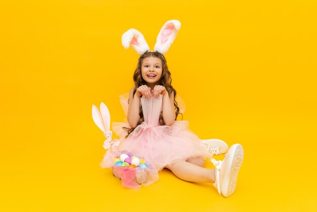 Pâques festive Une petite fille représente un lapin de Pâques avec un panier d'oeufs colorés Un enfant avec des oreilles de lapin sur un fond isolé jaune
