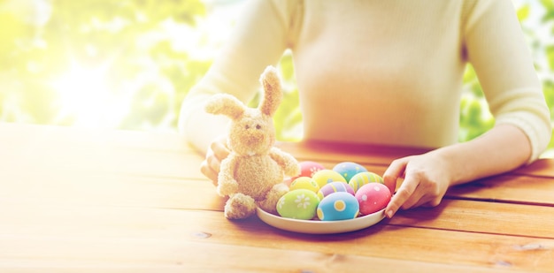 pâques, vacances, tradition et concept de personnes - gros plan sur des mains de femme avec des oeufs de pâques colorés sur une assiette et un lapin