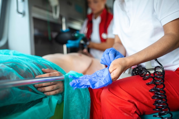 Un paramédic afro-américain met des gants en caoutchouc chirurgicaux bleus dans un véhicule ambulancier avec un technicien médical d'urgence blessé qui vérifie les signes vitaux de la victime