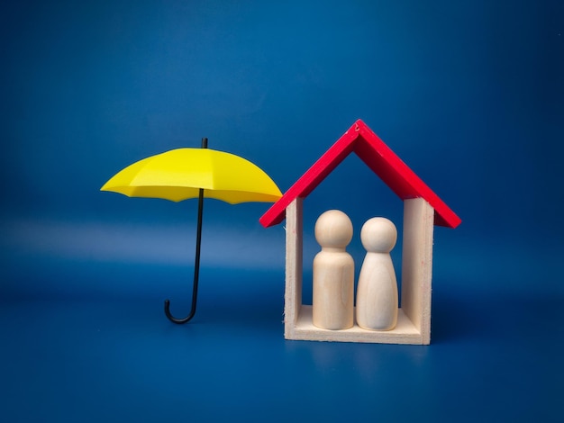 Un parapluie jaune protège la maison en bois et la poupée à chevilles sur fond bleu Planifier sauver les familles prévenir les risques et les crises concepts de soins de santé et d'assurance
