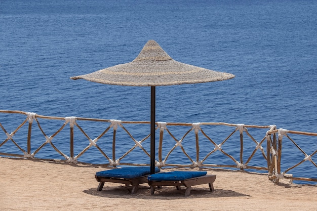 Parasols en paille avec chaises longues en bois à côté de l'eau de mer rouge sur la plage de sable du complexe à Sharm El Sheikh, Egypte