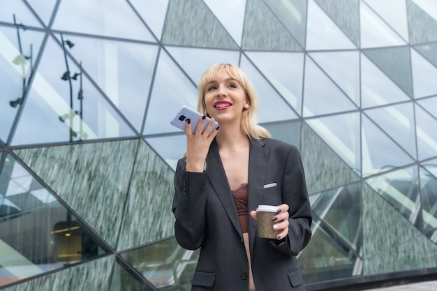 Parc D'affaires De Fille Entrepreneuriale Dans Un Immeuble Moderne Avec Verre Coloré, Jeune Blonde En Costume Envoyant Une Note Vocale Avec Un Café à Emporter