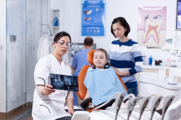 Parent et enfant dans le bureau de dentisterie regardant la radiographie dentaire détenue par le dentiste. Stomatologue expliquant le diagnostic des dents à la mère de l'enfant dans une clinique de santé tenant une radiographie.