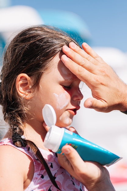 Un parent met de la crème solaire sur le visage d'une fille depuis un tube bleu au bord de la piscine d'un hôtel familial